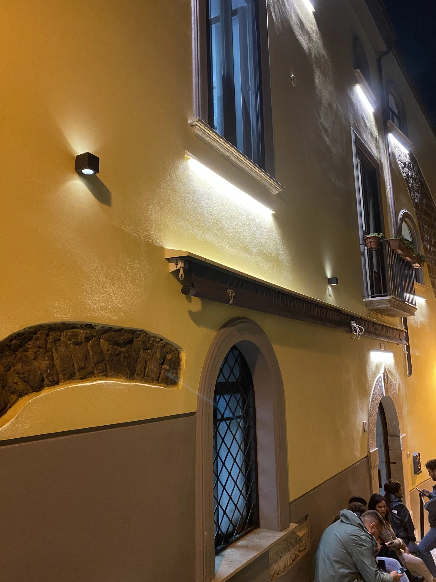 Facciata storica illuminata di notte a Pepe in Grani Caiazzo con applique LED architetturali e clienti in attesa nel vicolo